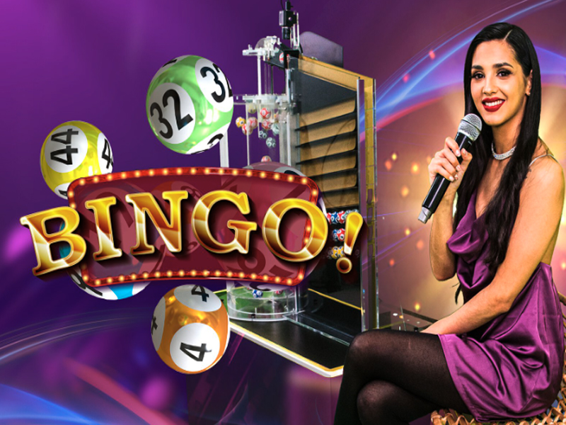 A female host in a purple dress holding a microphone, seated next to a bingo machine, with colorful bingo balls and a 'Bingo!' sign prominently displayed.