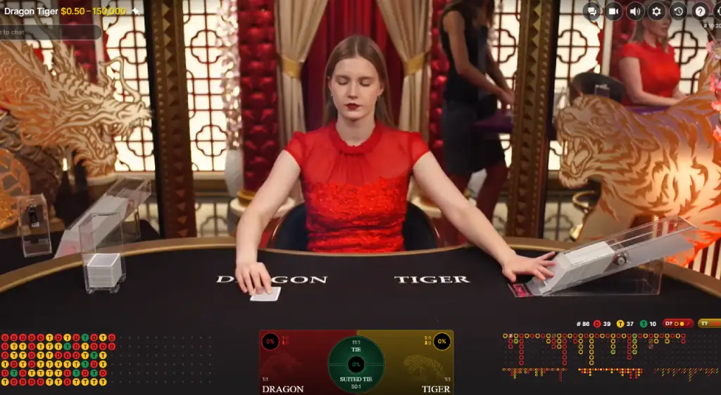 A live casino game of Dragon Tiger is shown, featuring a dealer in a red dress seated at a black gaming table with "Dragon" and "Tiger" betting areas clearly marked. The screen displays betting options, a live card dealing area, and game statistics. Behind the dealer, red and gold Asian-themed decor is visible, including dragon and tiger motifs.